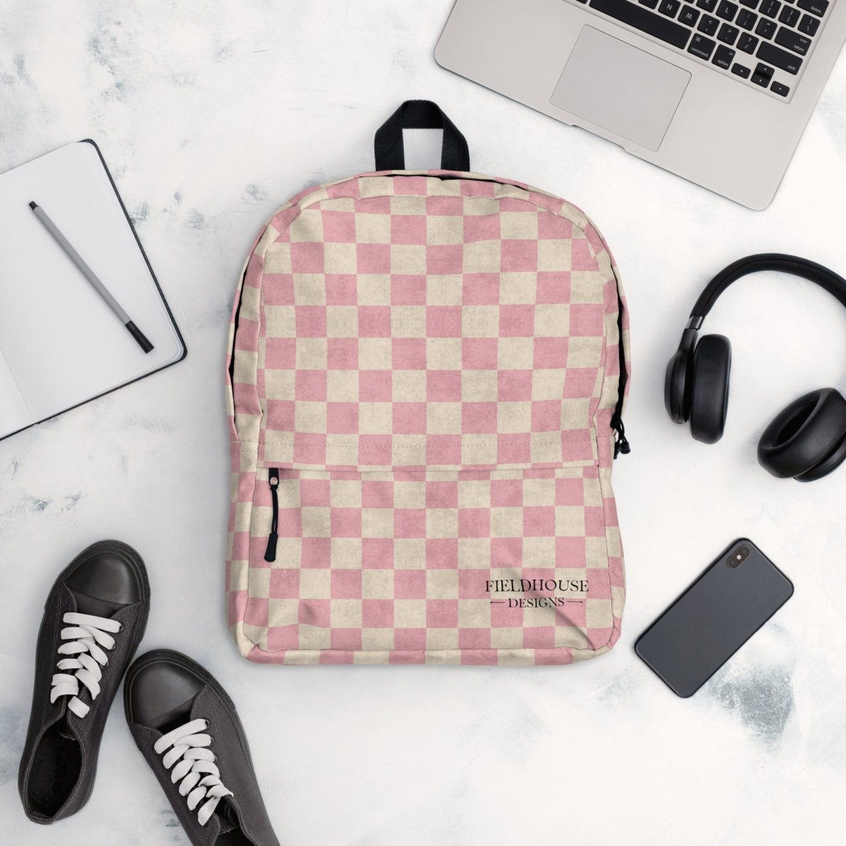 Pink and beige checkered backpack on a marble surface with a laptop, headphones, and shoes.