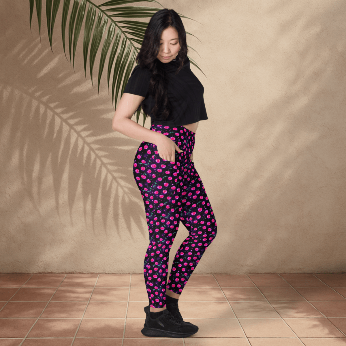 Woman wearing black crop top and pink cherry print leggings standing against a beige wall with a plant leaf.