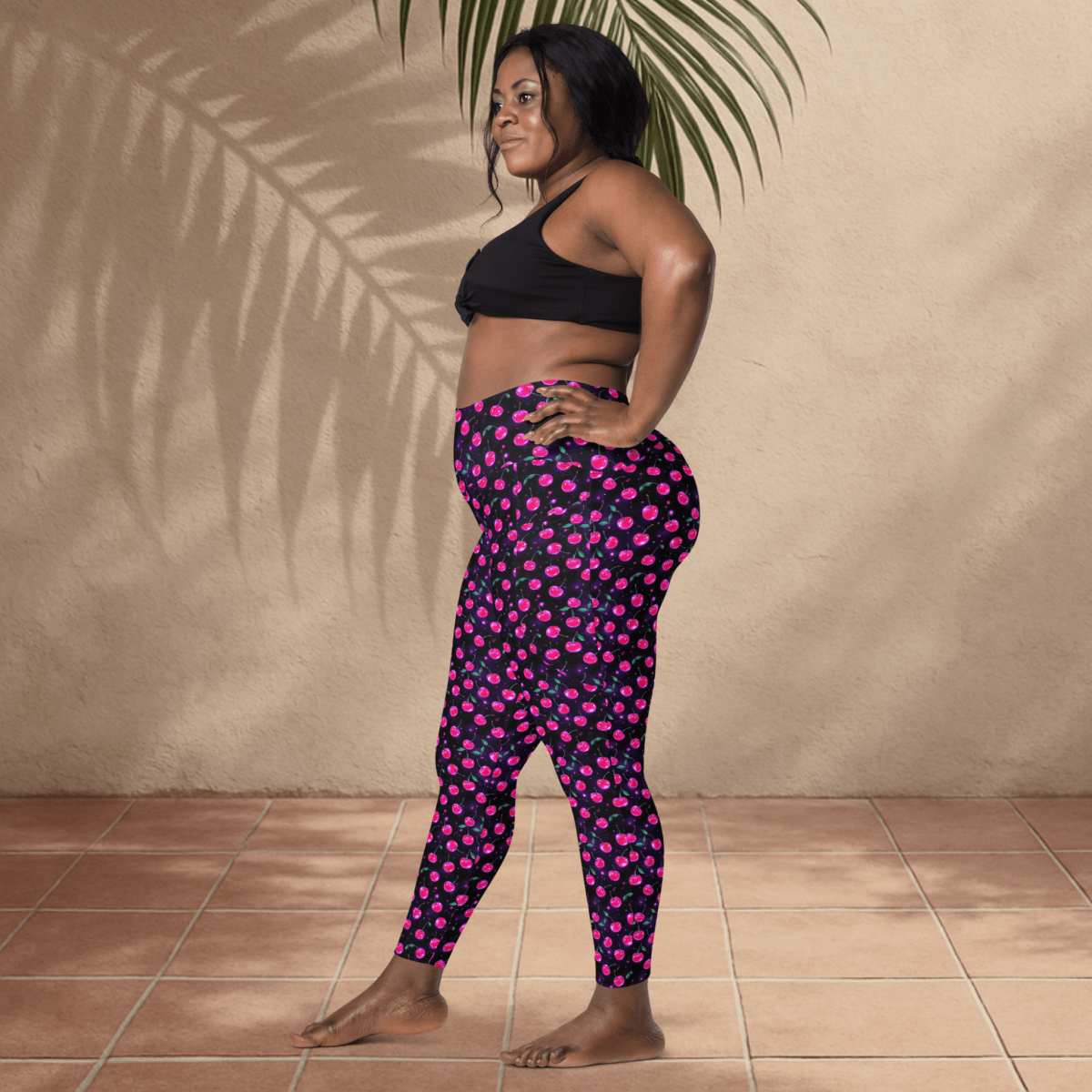 Woman wearing black sports bra and pink cherry print on black leggings standing against a beige wall.
