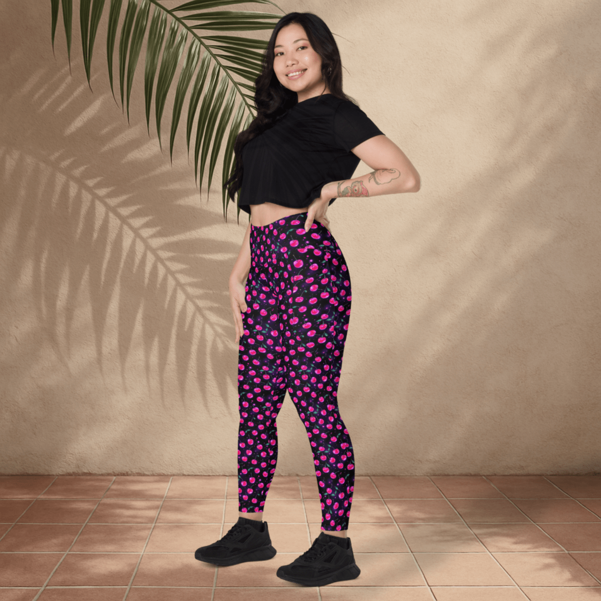 Woman wearing black crop top and pink cherry print on black pants standing against a beige wall with a palm leaf.