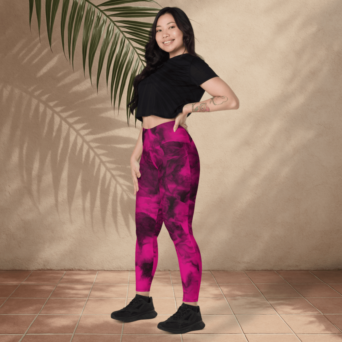 Woman wearing pink and black patterned leggings and black top standing against a beige wall with a plant.