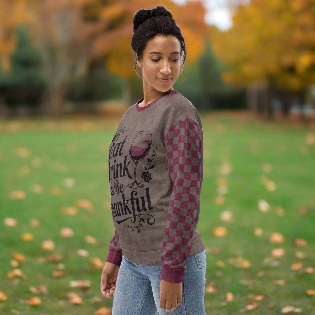 Woman wearing a sweater with 'Eat Drink and be Thankful' text and wine glass design in an outdoor setting with trees and grass.