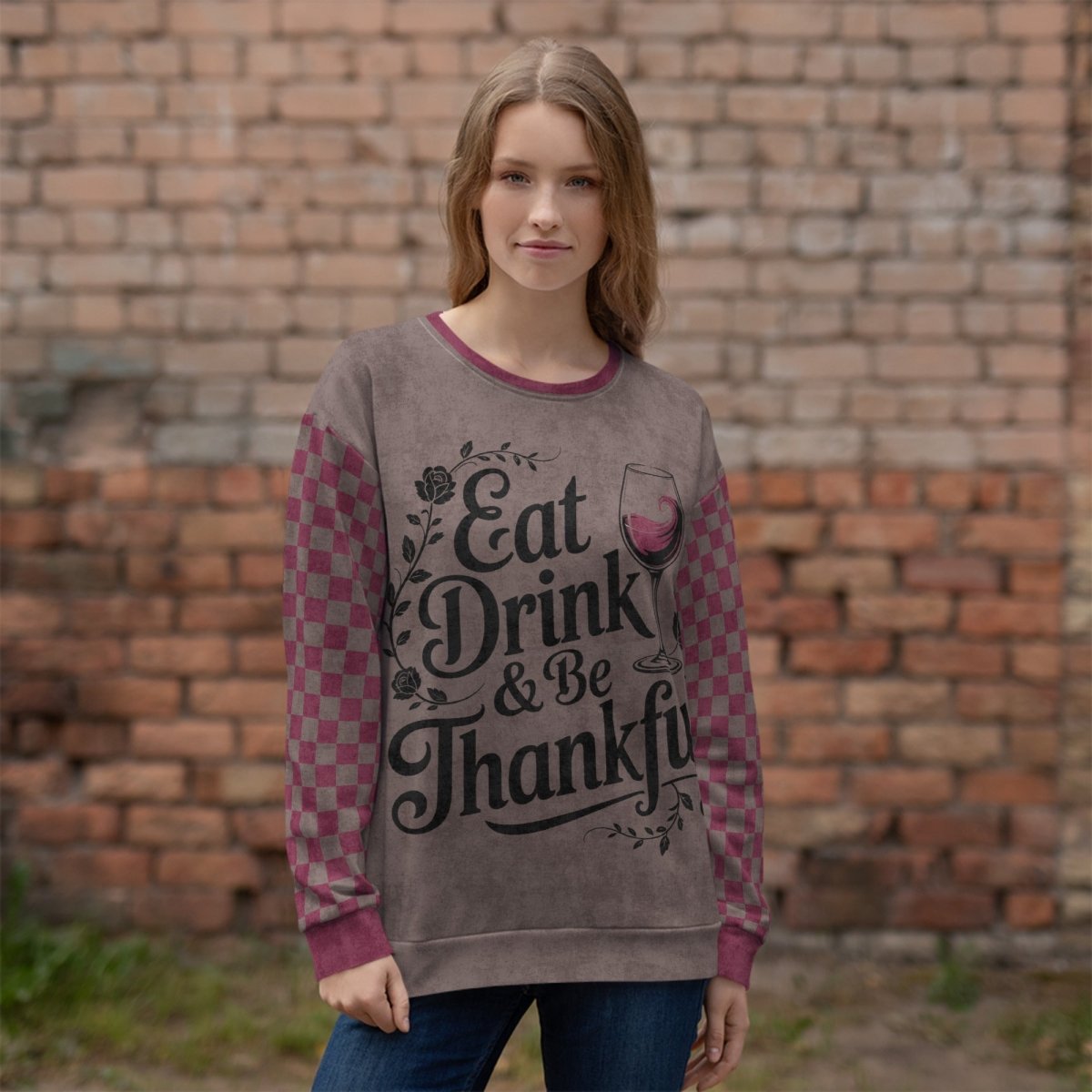 Woman wearing a sweater with 'Eat Drink & Be Thankful' text against a brick wall.