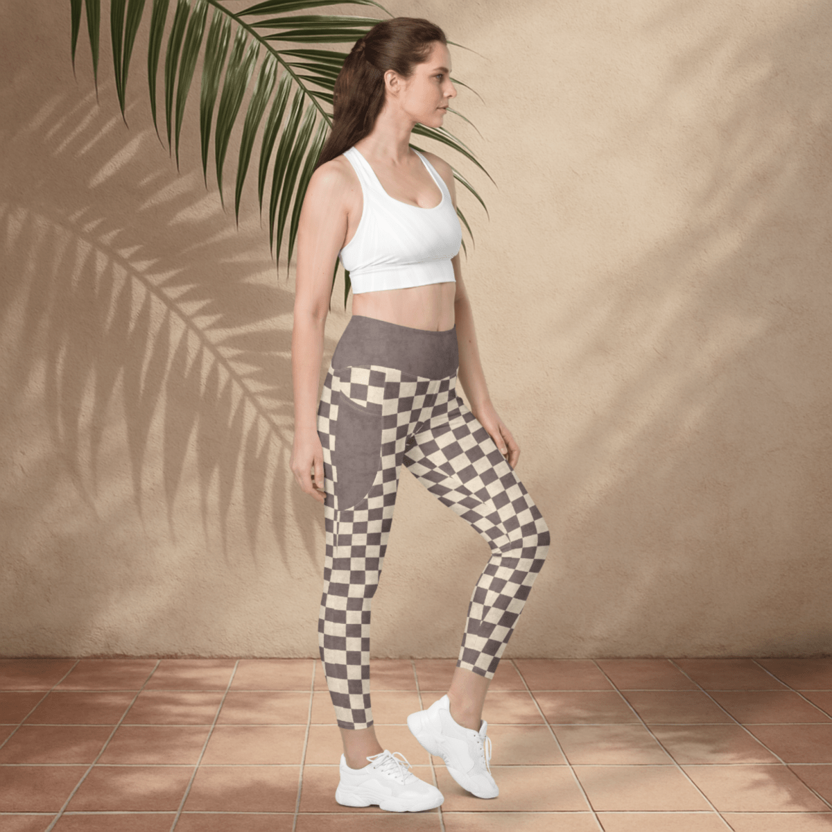 Woman wearing a white sports bra and checkered leggings standing against a beige wall with a palm leaf.