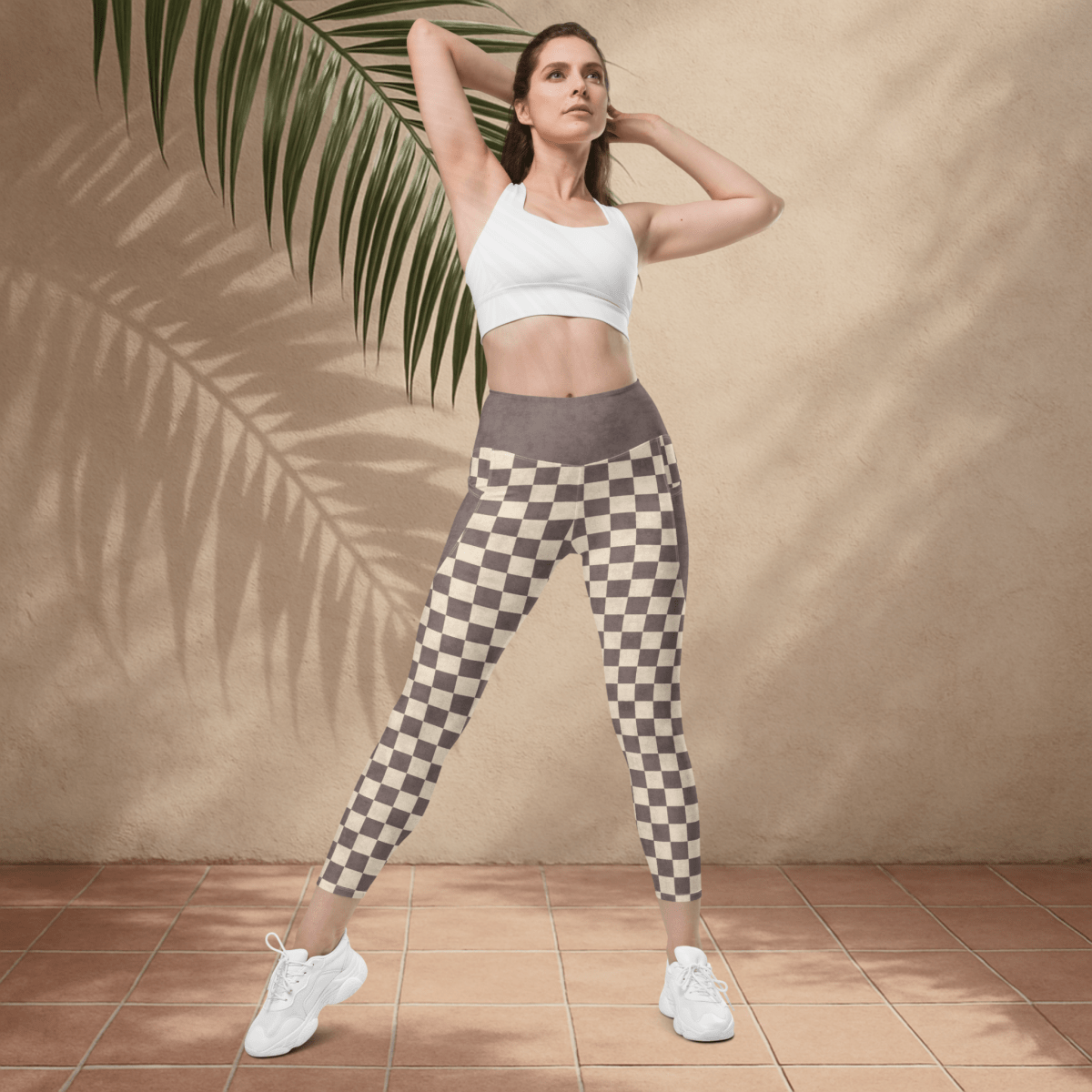Woman wearing a white sports bra and checkered leggings standing against a beige wall with a palm leaf.