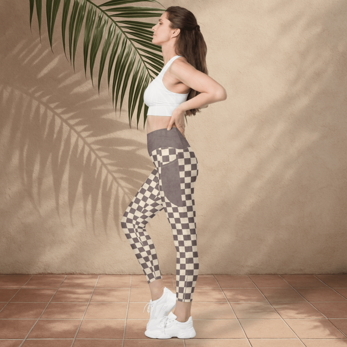 Woman wearing checkered leggings and a white sports bra against a beige wall with a palm leaf.