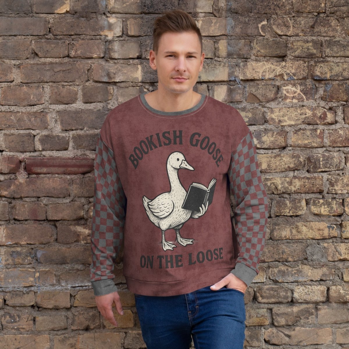 Man wearing a burgundy checkered sweater with a graphic of a goose holding a book and text 'Bookish Goose on the Loose' against a stone wall.