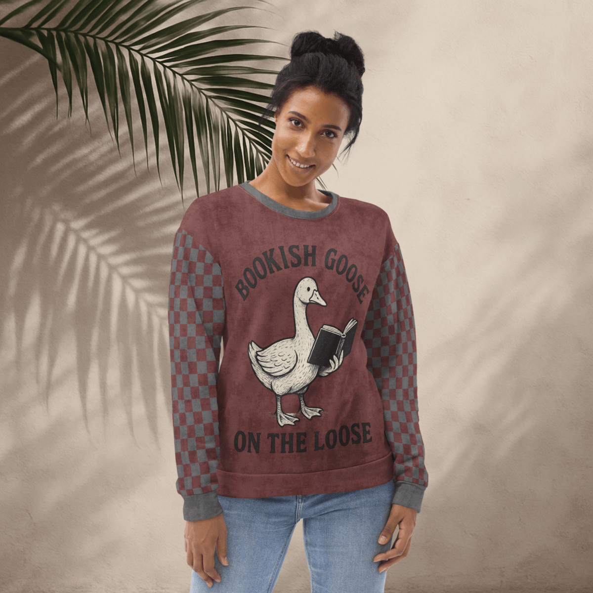 Woman wearing a maroon sweatshirt with a graphic of a goose holding a book and text 'Bookish Goose on the Loose', standing against a beige wall with a palm leaf.