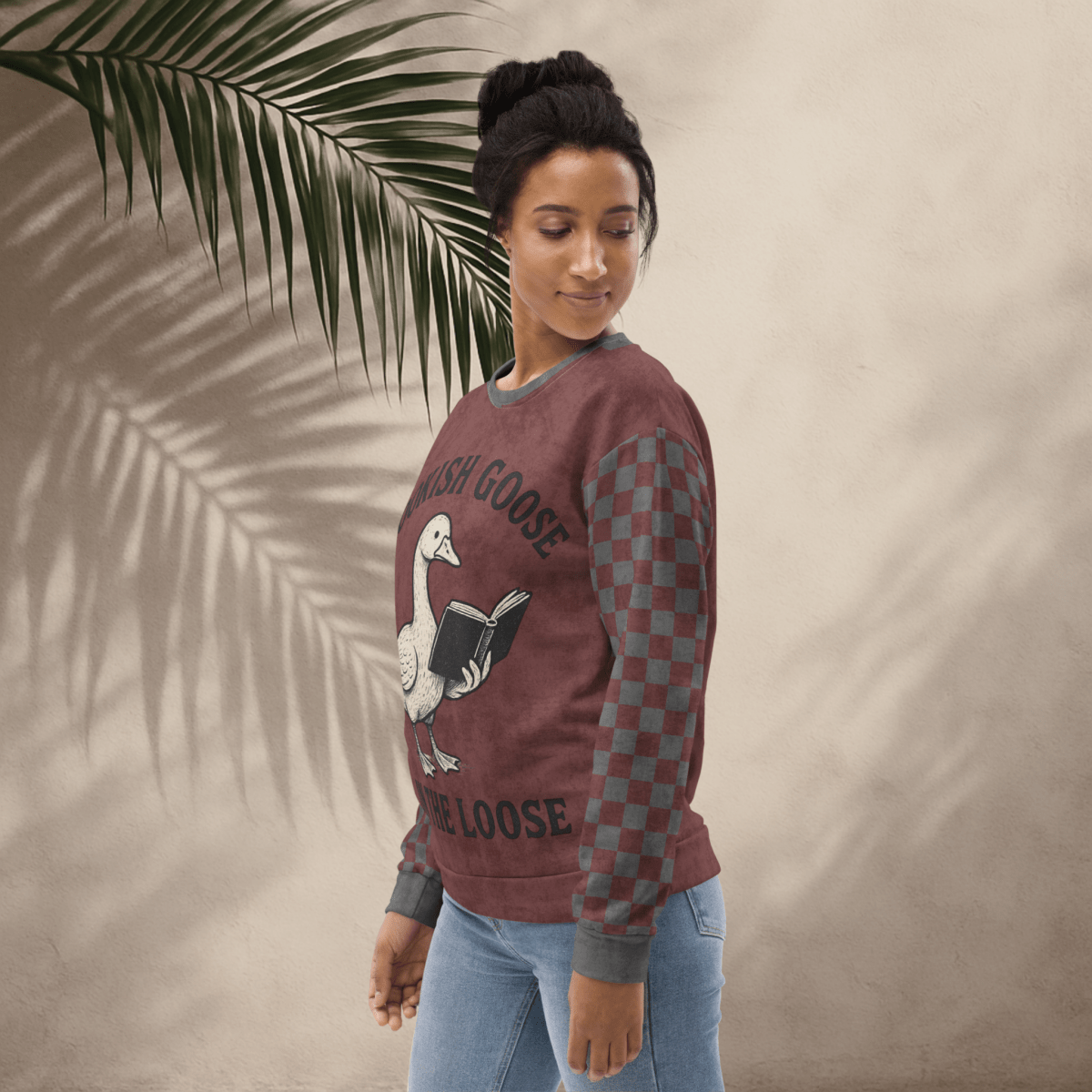 Woman wearing a burgundy crewneck sweater with a graphic design, standing against a beige wall with a palm leaf.