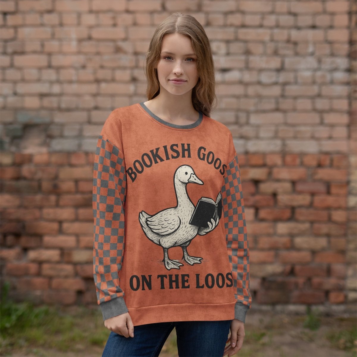 Person wearing a sweater with a goose reading a book and text 'Bookish Goose on the Loose" in front of a brick wall