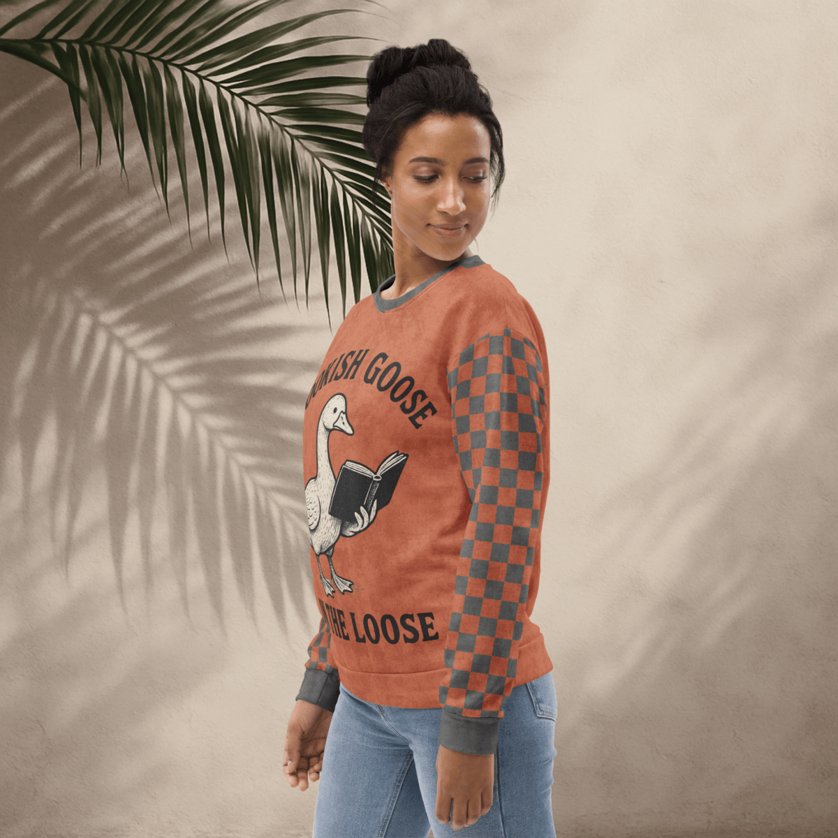Woman wearing a terracotta checkered long-sleeve shirt with a goose reading a book graphic and text 'Bookish Goose on the Loose', standing against a beige wall with a palm leaf.