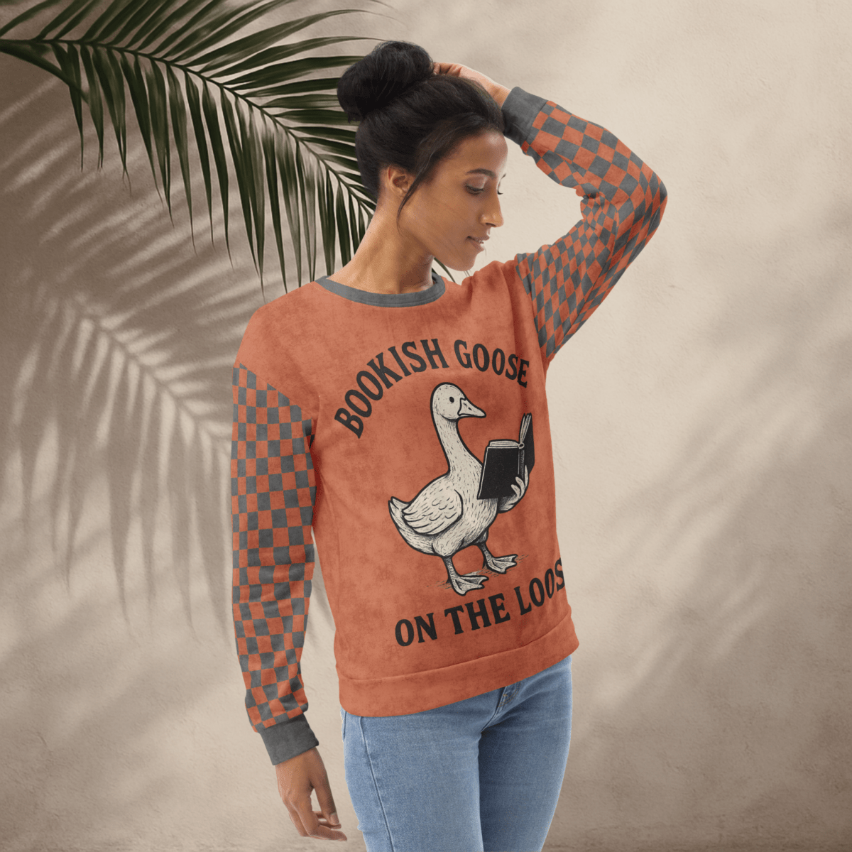 Woman wearing a sweater with a goose reading a book graphic and text 'Bookish Goose on the Loose', standing against a neutral background with a leaf.