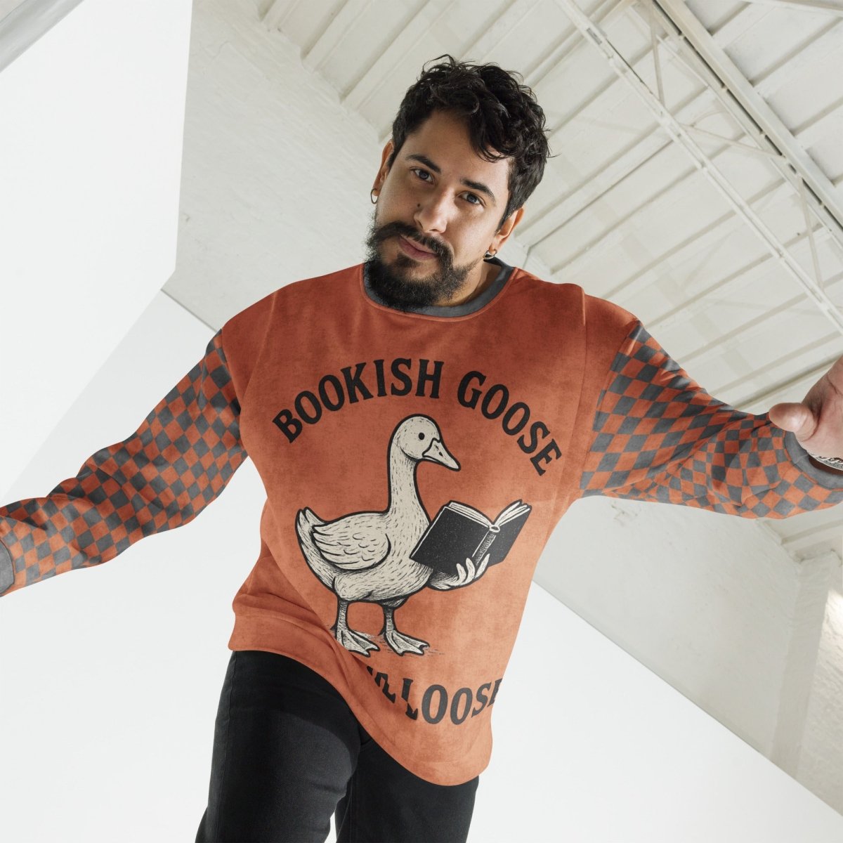 Man wearing a terracotta checkered 'Bookish Goose' t-shirt with a graphic of a goose holding a book, standing in a modern indoor setting.