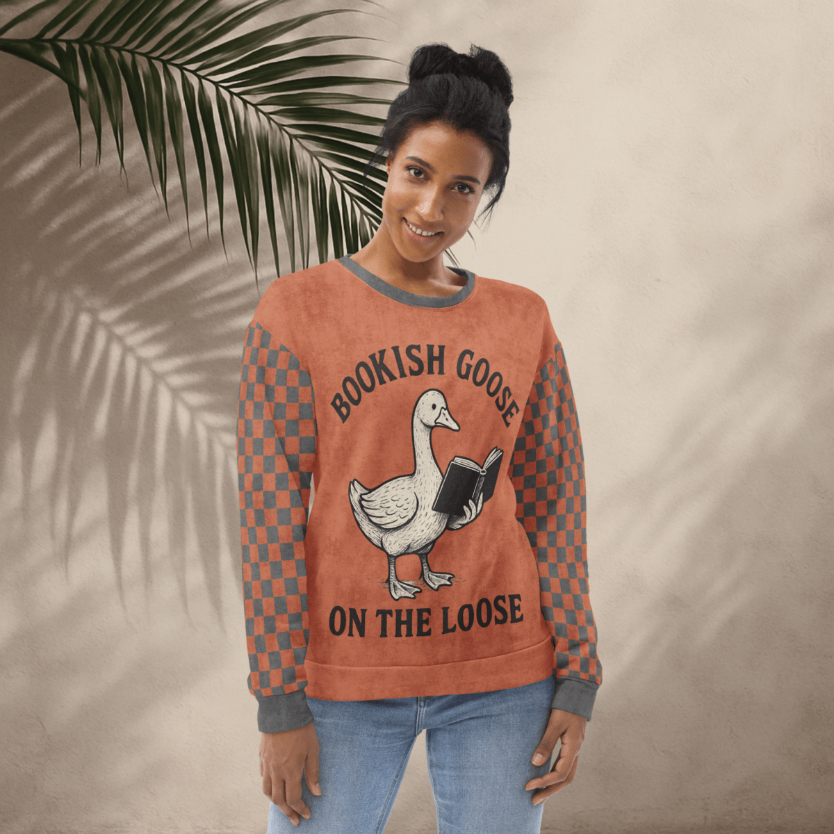 Woman wearing a sweater with a goose reading a book graphic and text 'Bookish Goose on the Loose', standing against a beige wall with a leafy branch.