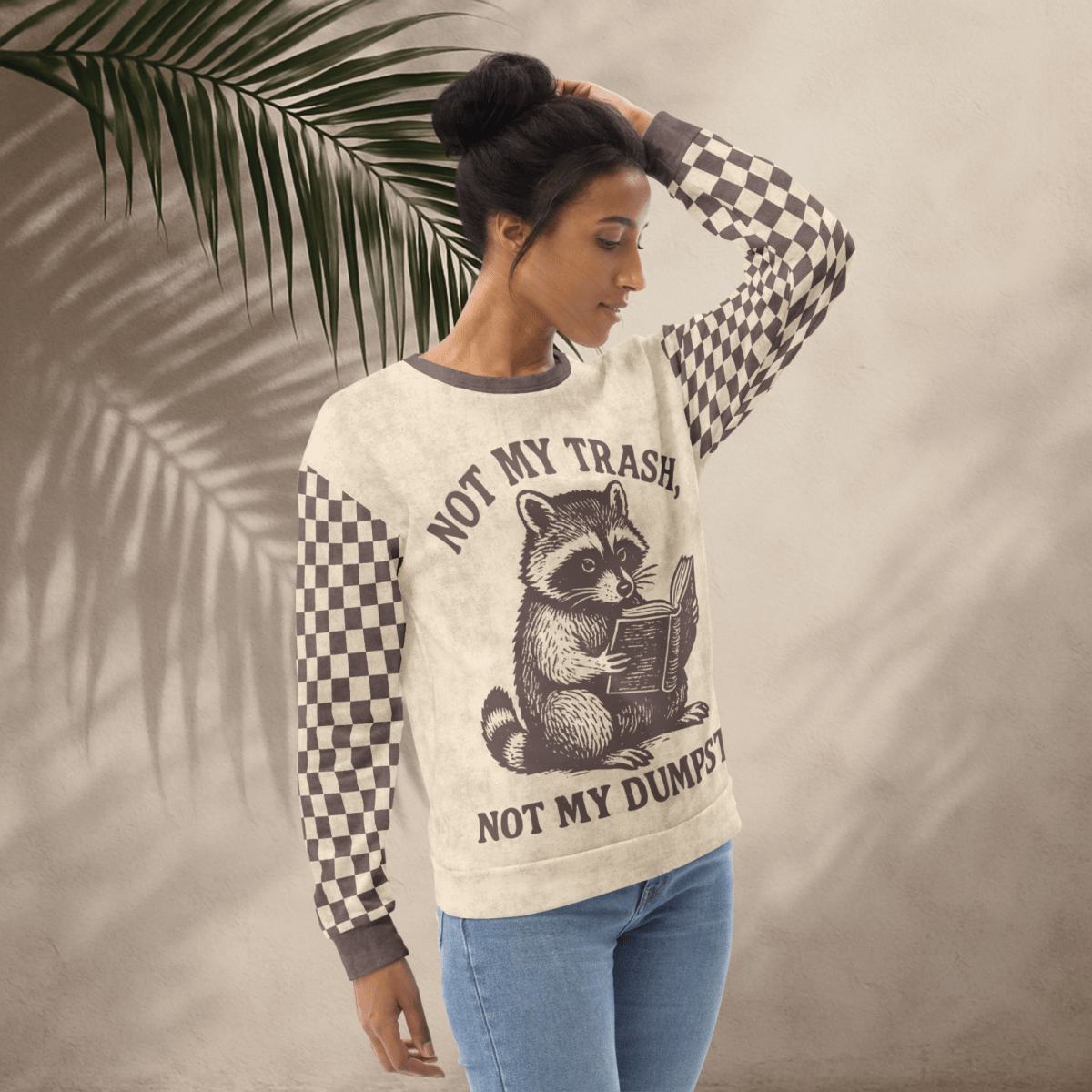 model wearing a Beige crewneck sweatshirt with espresso checkered sleeves. Featuring a raccoon reading a book graphic and text 'Not my trash, not my dumpster'