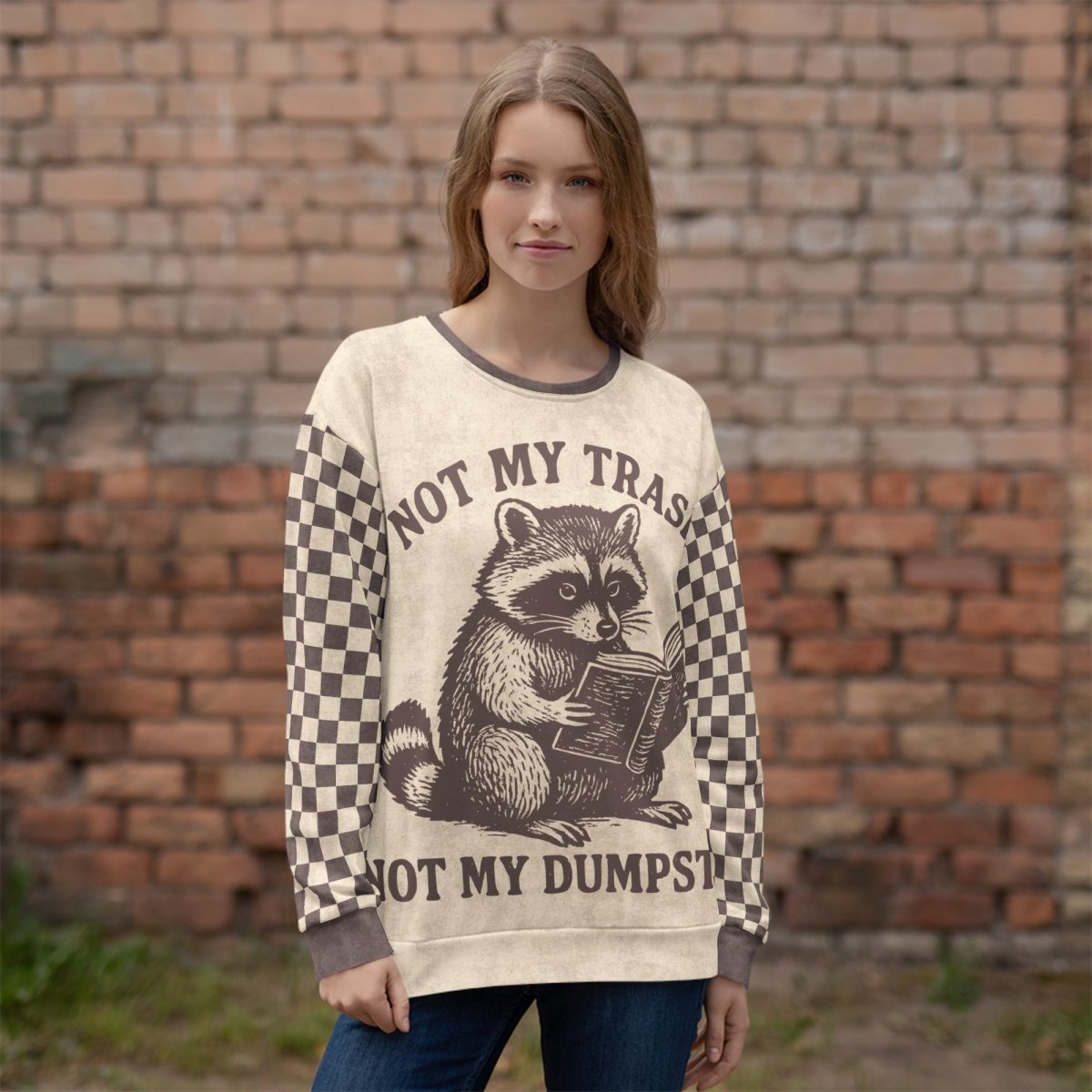Woman wearing a sweater with a raccoon reading a book graphic and text "Not my Trash, Not my Dumpster" in front of a brick wall.