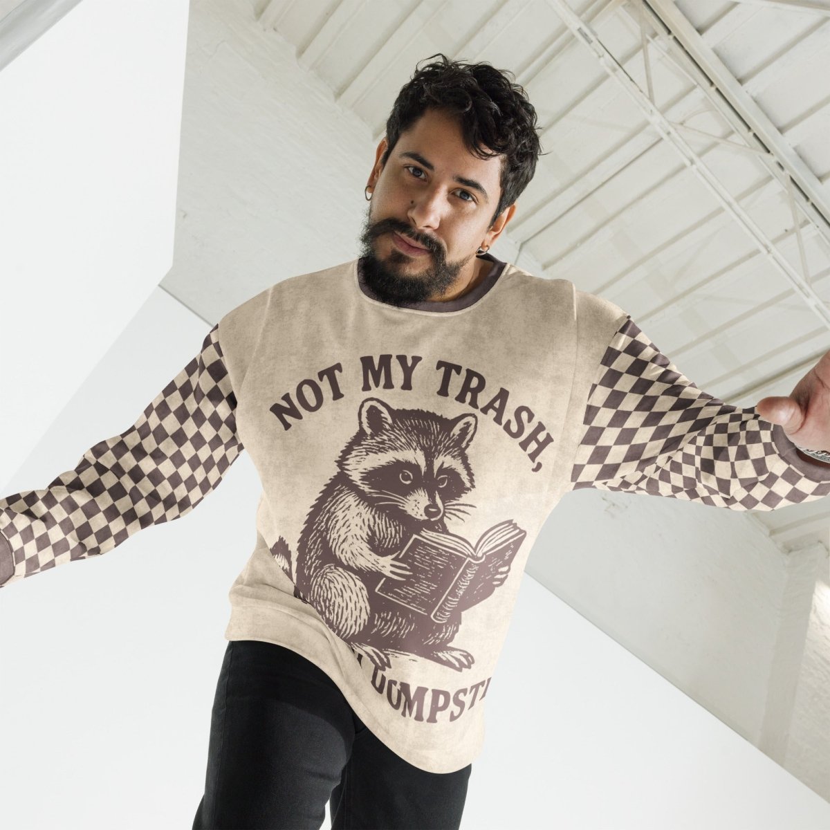 Man wearing a Beige crewneck sweatshirt with espresso checkered sleeves. Featuring a raccoon reading a book graphic and text 'Not my trash, not my dumpster'