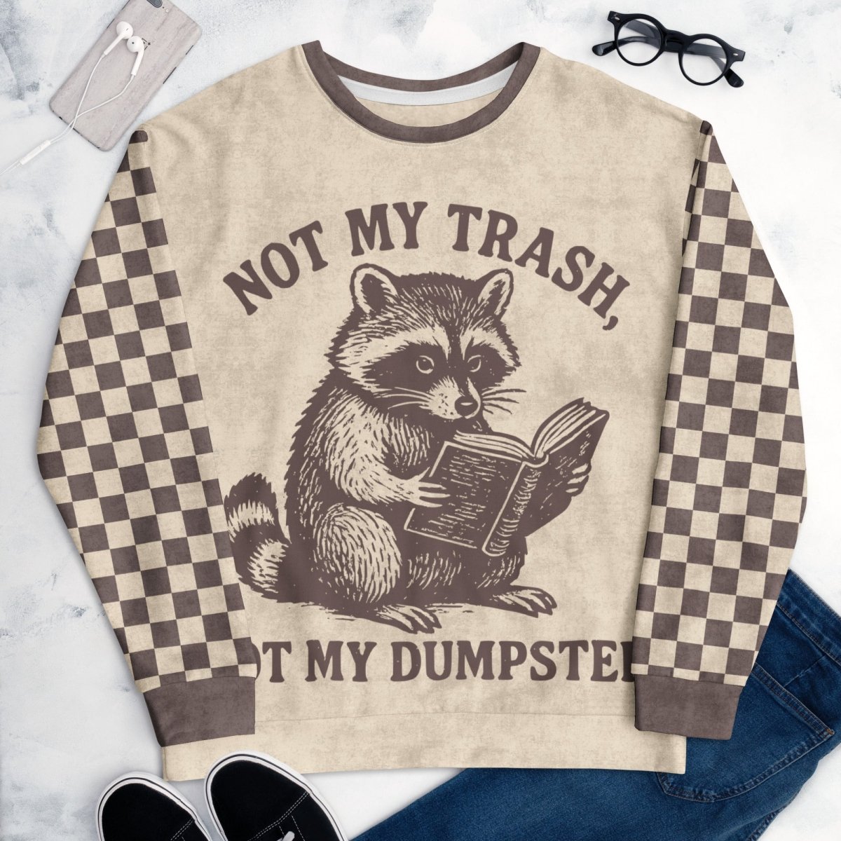 Beige crewneck sweatshirt with espresso checkered sleeves. Featuring a raccoon reading a book graphic and text 'Not my trash, not my dumpster' on a marble surface.