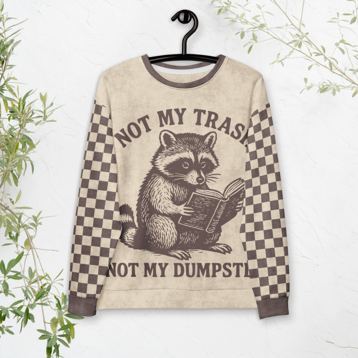 hanging Beige crewneck sweatshirt with espresso checkered sleeves. Featuring a raccoon reading a book graphic and text 'Not my trash, not my dumpster'