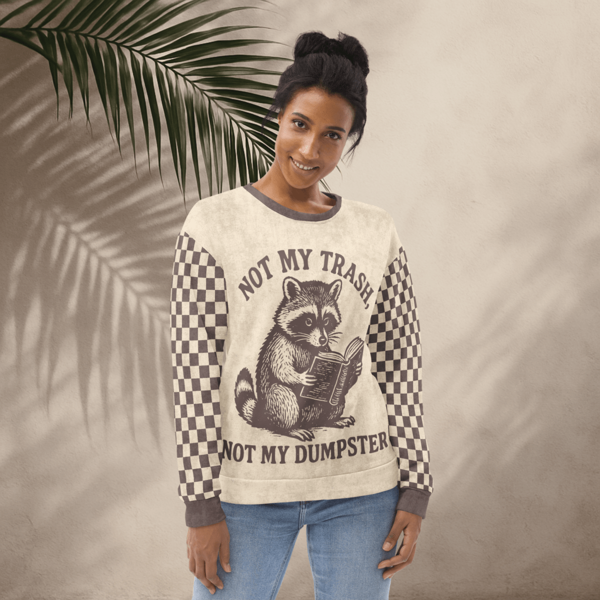 Beige crewneck sweatshirt with espresso checkered sleeves. Featuring a raccoon reading a book graphic and text 'Not my trash, not my dumpster'