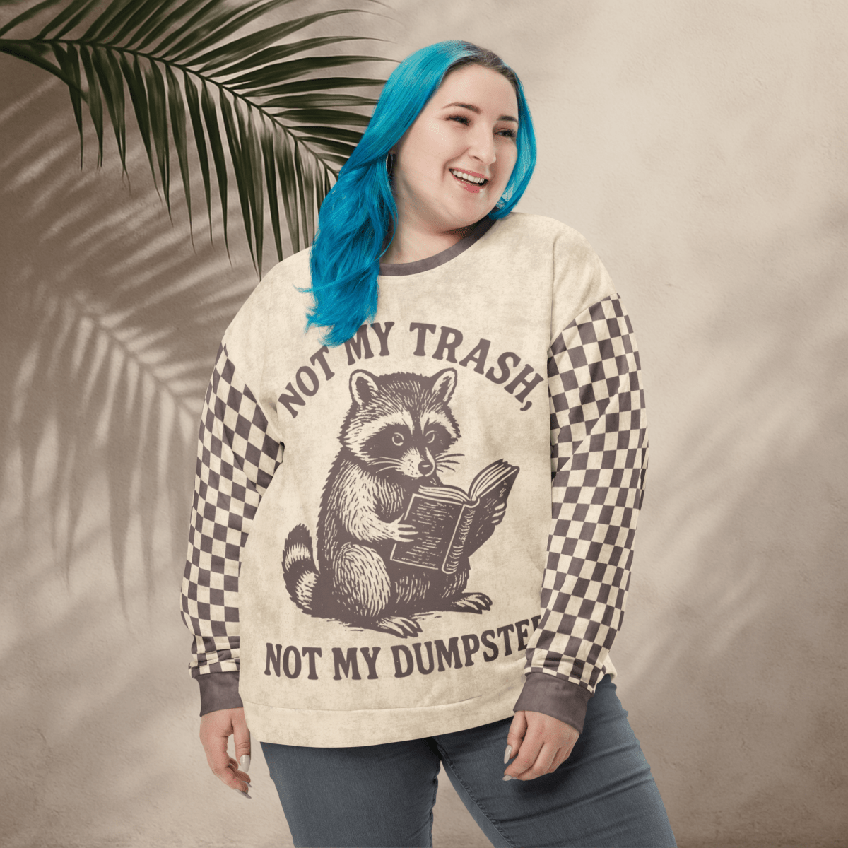 model wearing a Beige crewneck sweatshirt with espresso checkered sleeves. Featuring a raccoon reading a book graphic and text 'Not my trash, not my dumpster'