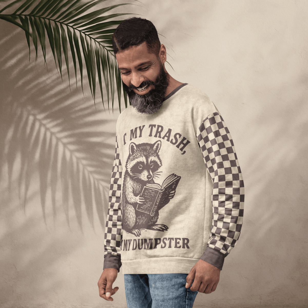model wearing a Beige crewneck sweatshirt with espresso checkered sleeves. Featuring a raccoon reading a book graphic and text 'Not my trash, not my dumpster'