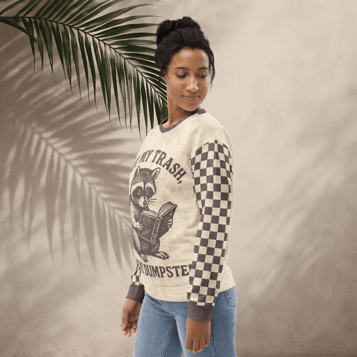 model wearing Beige crewneck sweatshirt with espresso checkered sleeves. Featuring a raccoon reading a book graphic and text 'Not my trash, not my dumpster'