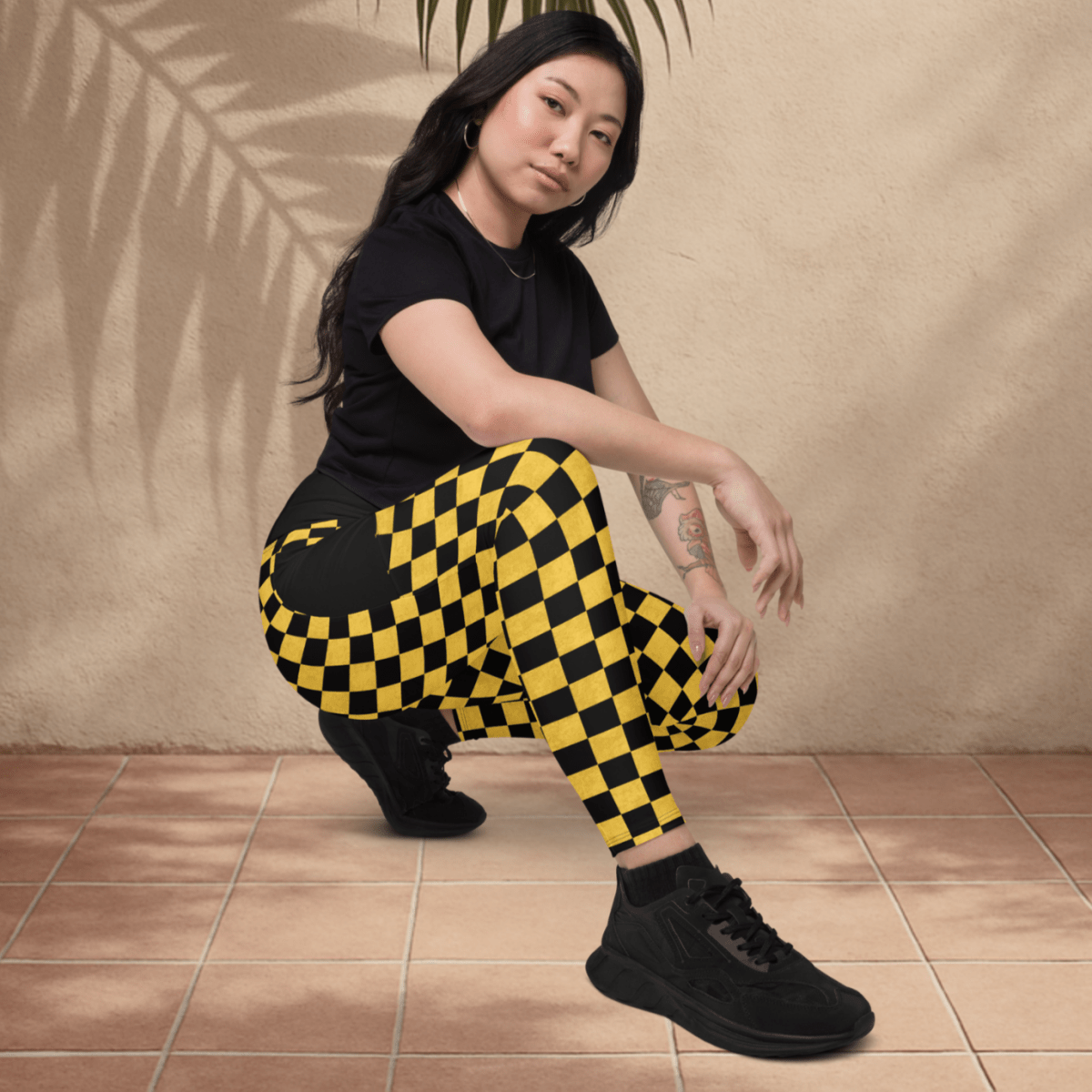 Person wearing a black t-shirt and yellow checkered pants posing against a beige wall with a palm leaf.