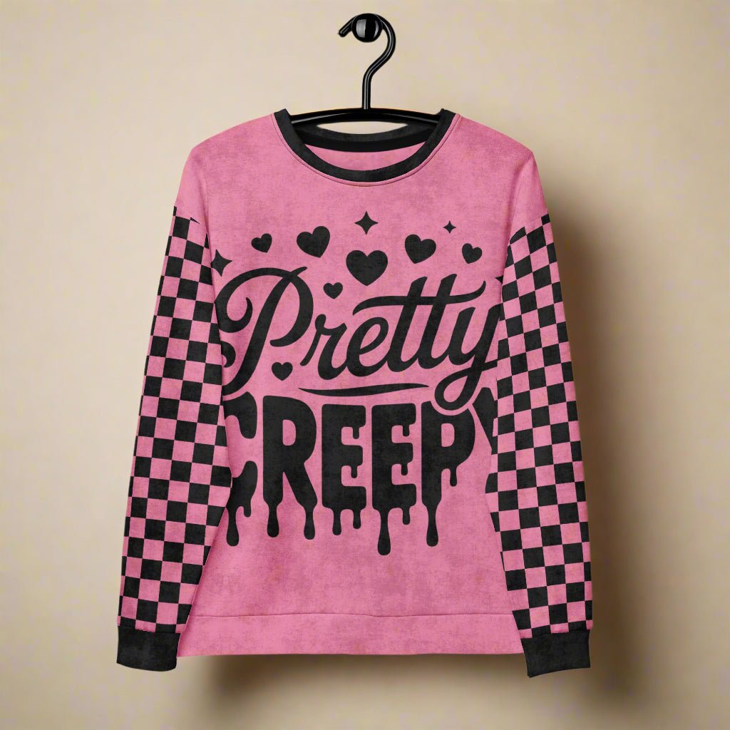 Pink sweatshirt with 'Pretty Creepy' text and checkered sleeves on a hanger against a beige background