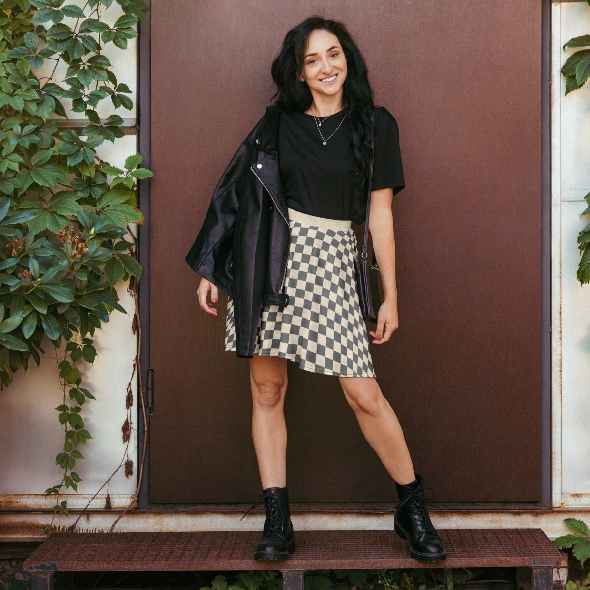 Woman standing in front of a brown door wearing a black top, checkered skirt, and black boots.