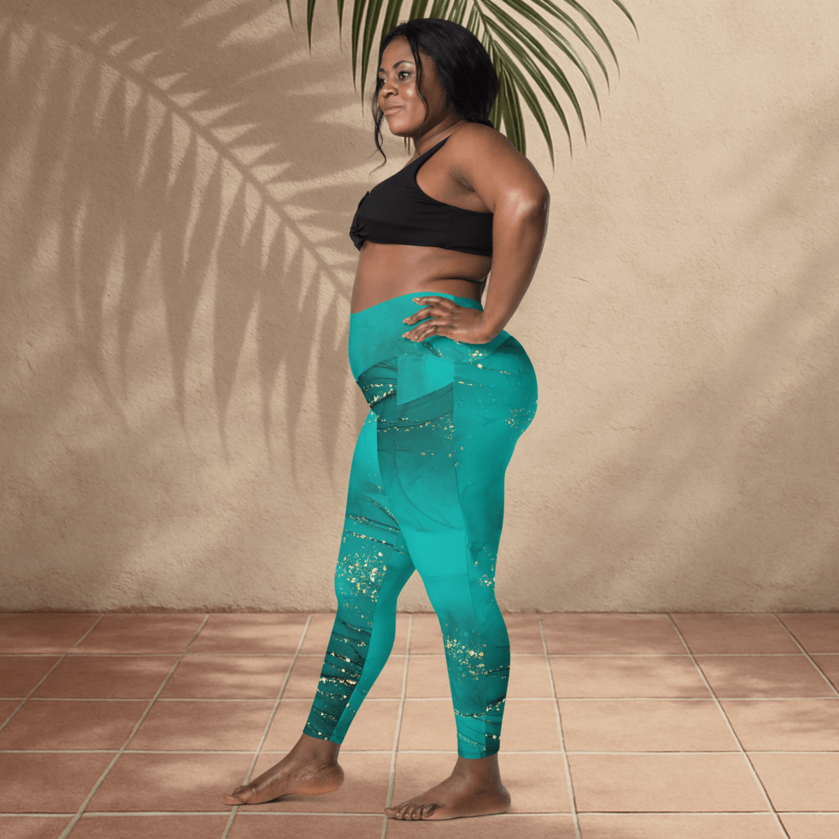 Woman wearing teal leggings and black sports bra standing against a beige wall with a plant.