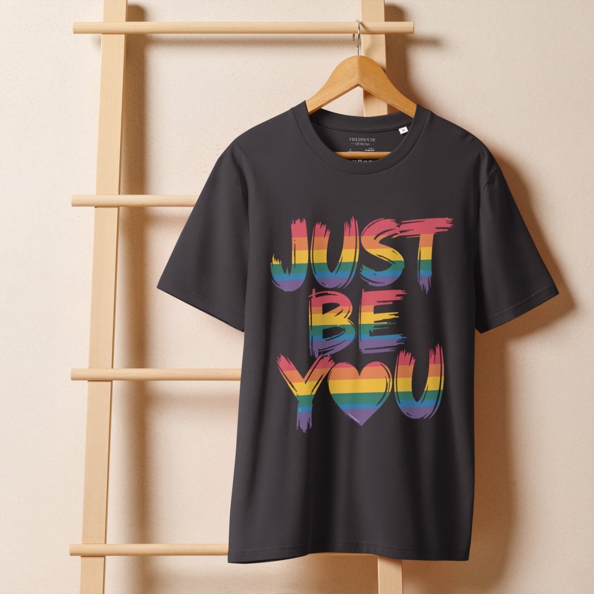 anthracite t-shirt with 'JUST BE YOU' in colorful letters on a wooden hanger against a beige wall.