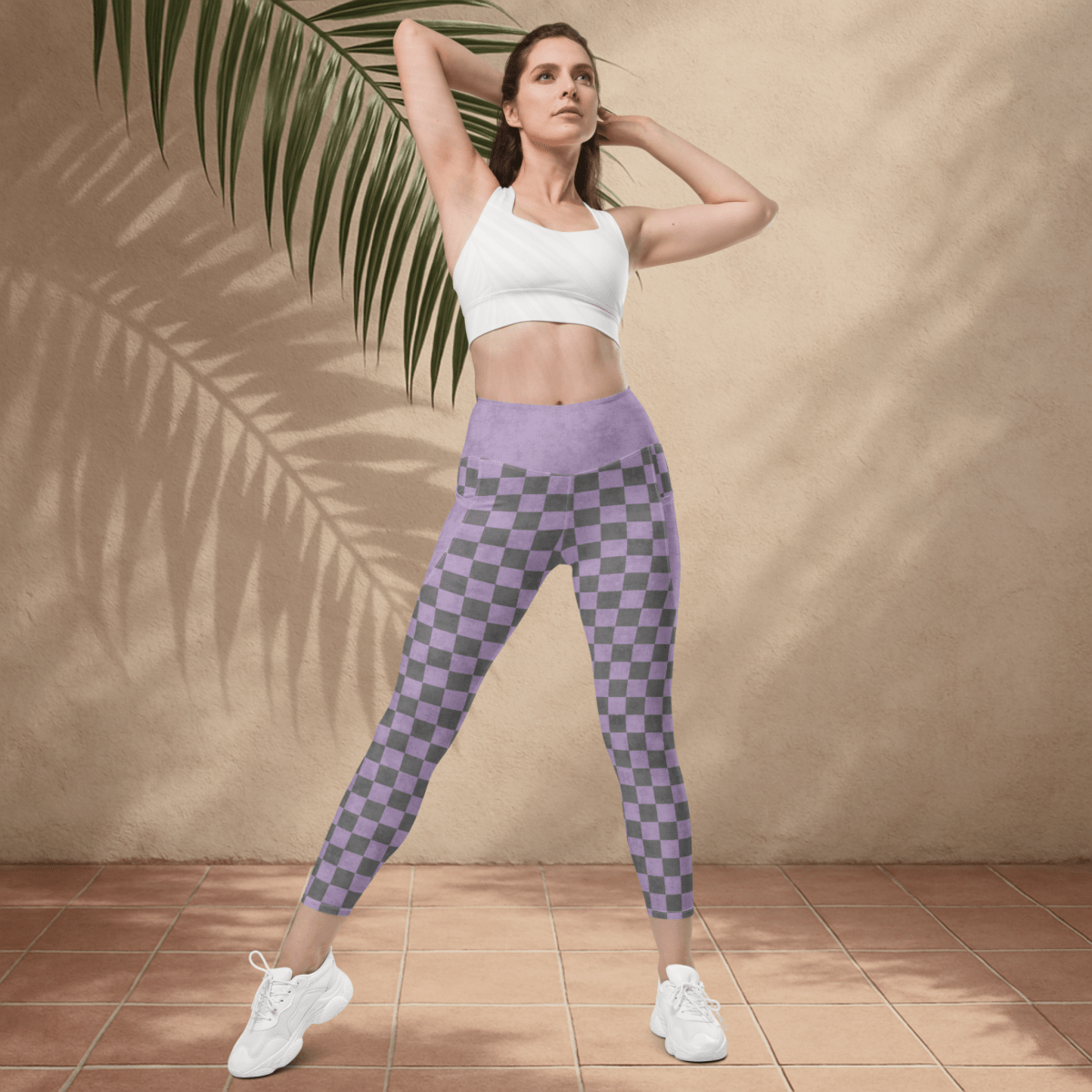 Woman wearing a white sports bra and purple checkered leggings standing against a beige wall with a palm leaf.