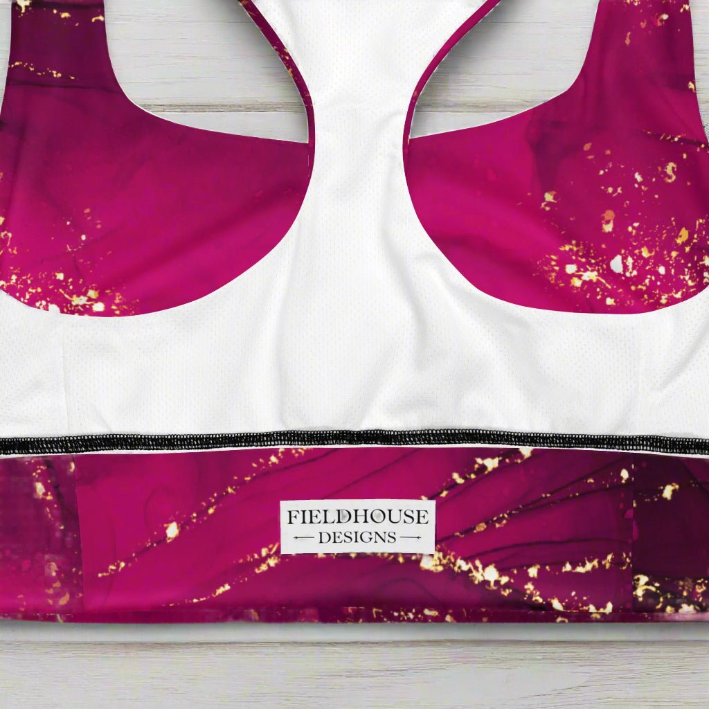 Pink sports bra with gold accents on a wood background, featuring the Fieldhouse Designs brand.
