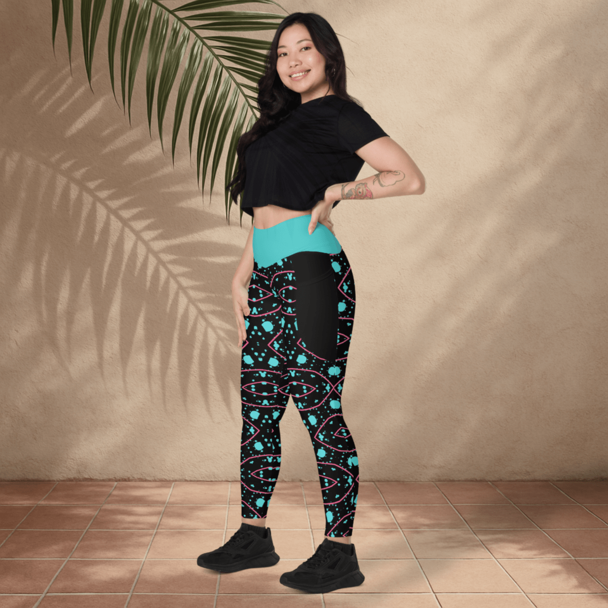 Woman wearing a black crop top and colorful patterned leggings standing against a beige wall with a plant.