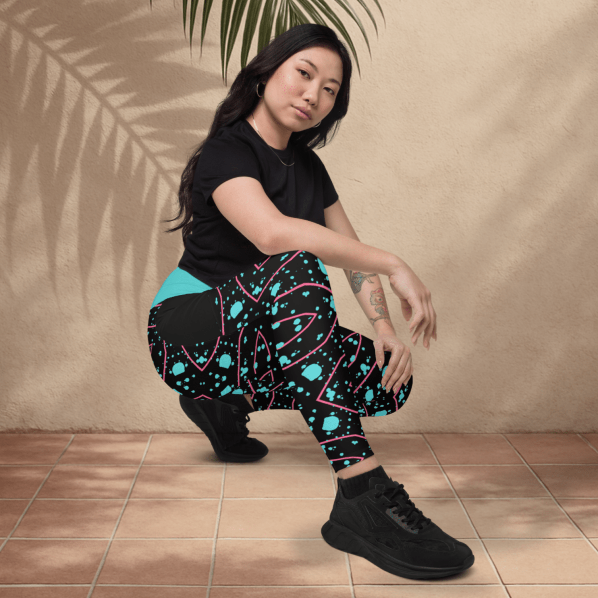 Woman wearing black and teal patterned pants sitting on a tiled floor with a beige wall and plant in the background.