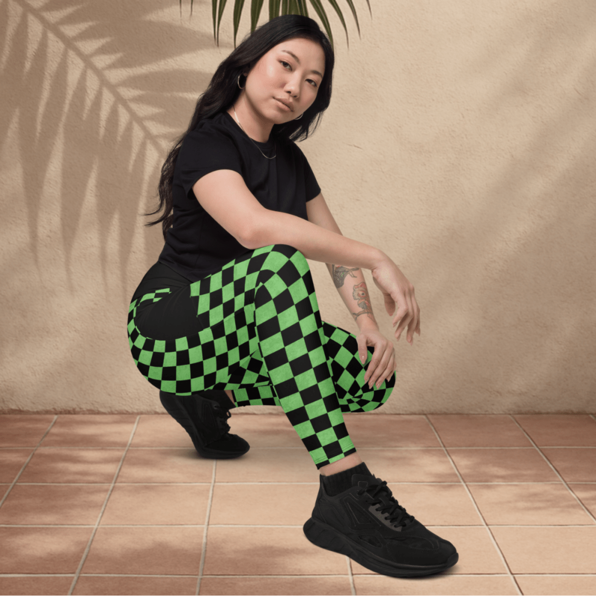 Person wearing black and green checkered pants with a leafy branch above them against a beige wall.
