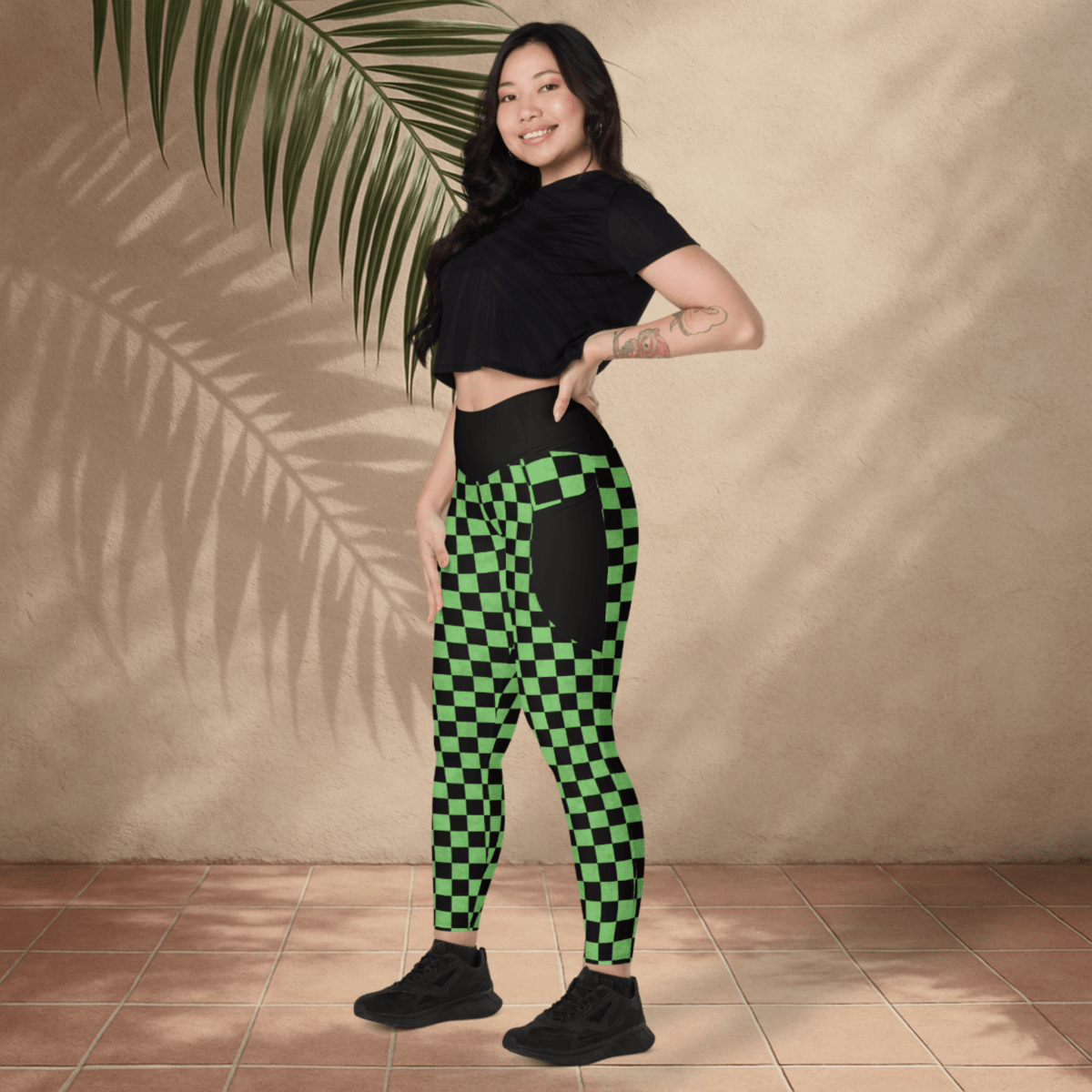 Woman wearing black crop top and green checkered pants standing against a beige wall with a palm leaf.