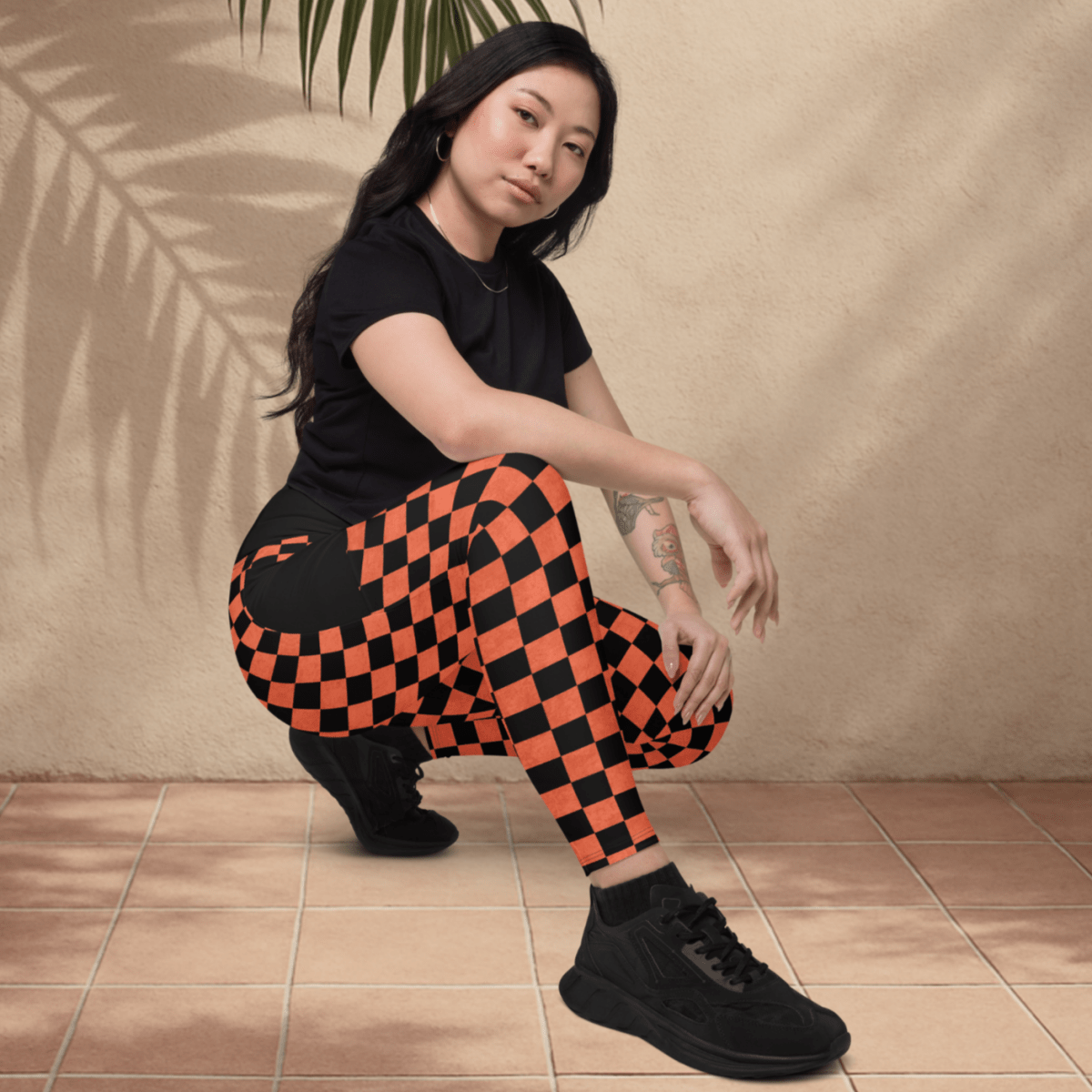 Person wearing black t-shirt and orange checkered pants posing against a beige wall with a palm leaf.
