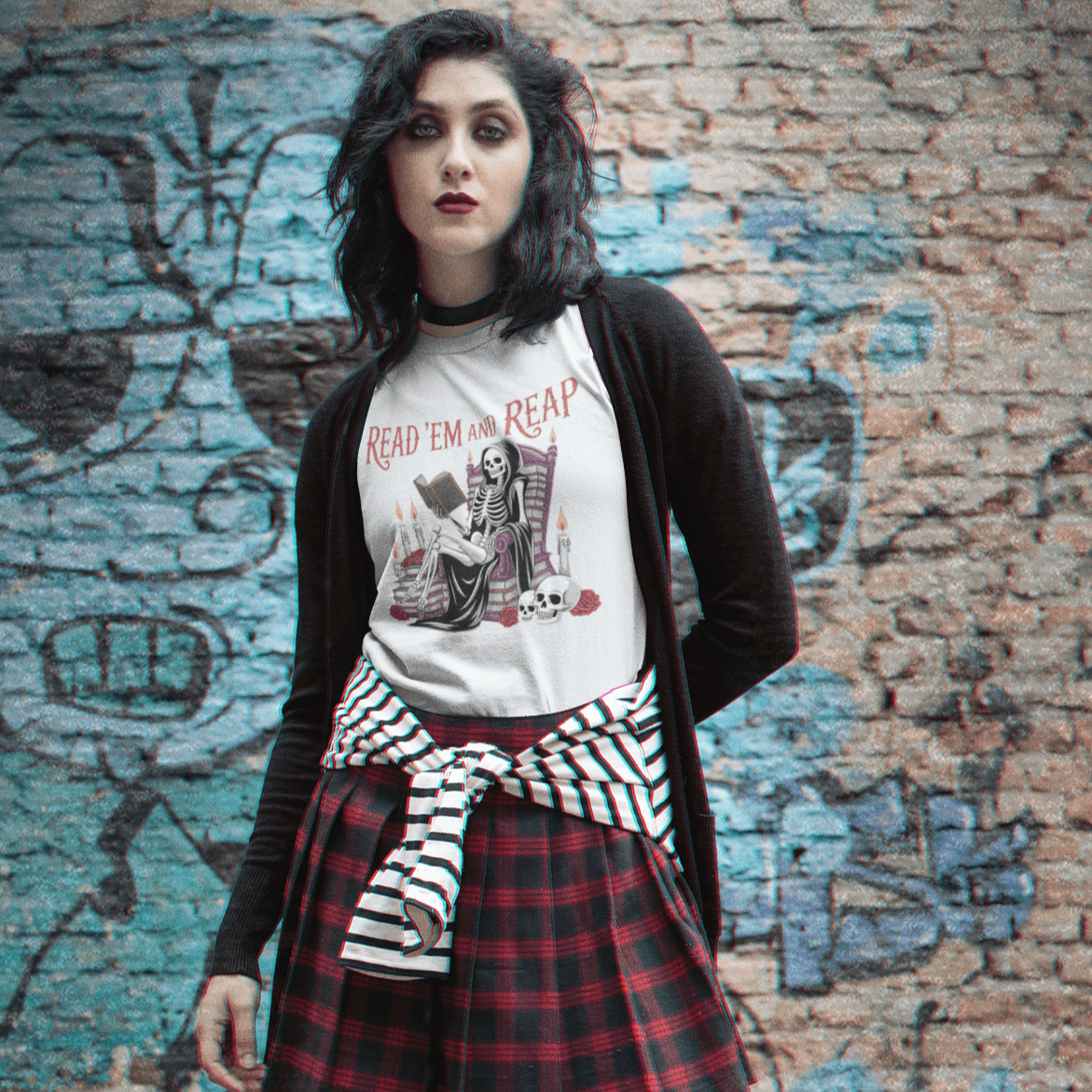 Person wearing a plaid skirt, graphic t-shirt, and black jacket in front of a graffiti-covered brick wall.