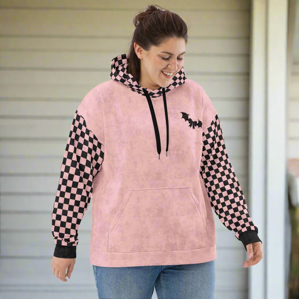 Person wearing a pink hoodie with checkered sleeves and a bat design, standing in front of a neutral background.