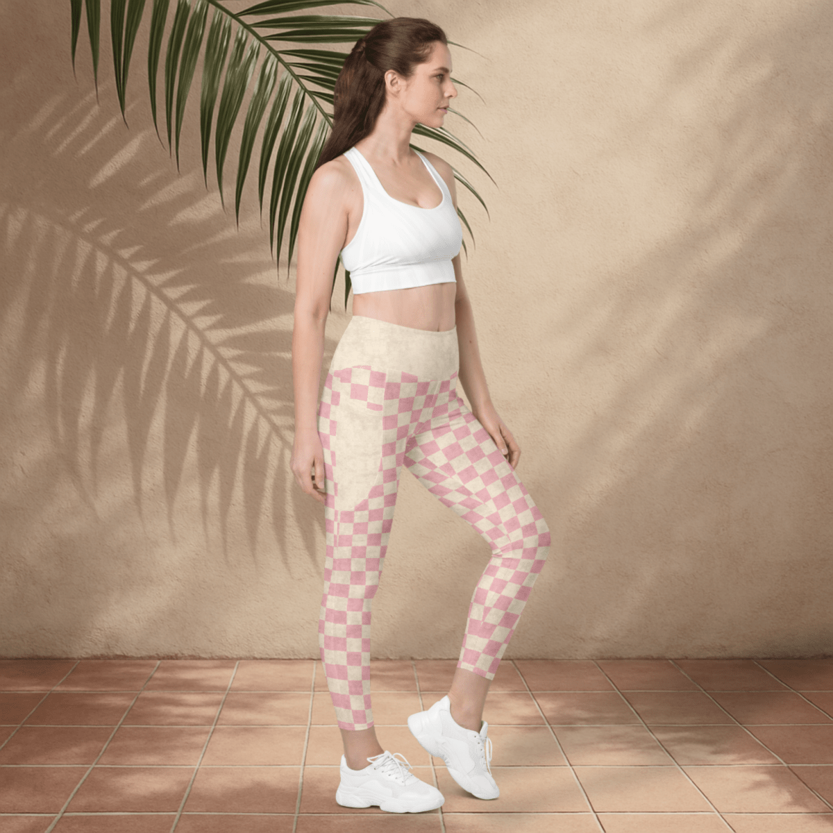 Woman wearing a white sports bra and pink checkered leggings standing against a beige wall with a palm leaf.