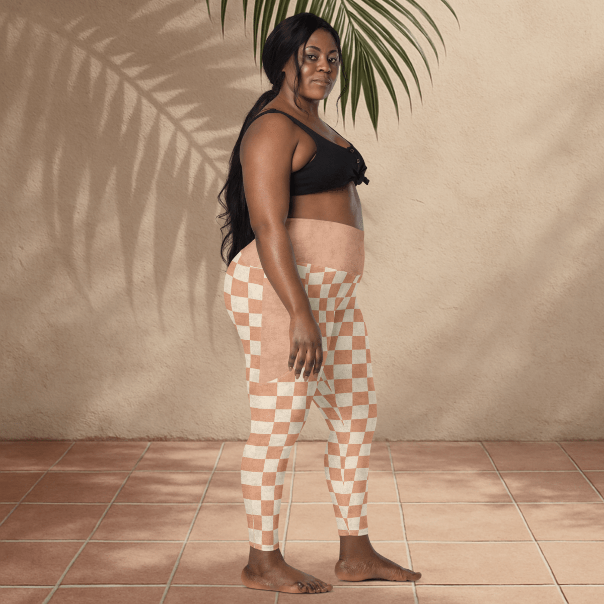 Woman wearing checkered pants standing against a beige wall with a plant.