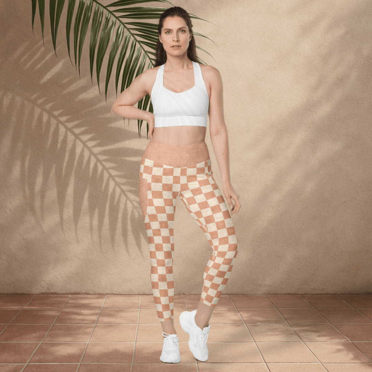 Woman wearing a white sports bra and checkered leggings standing against a beige wall with a plant.