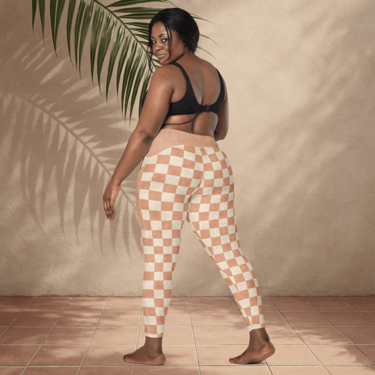 Woman wearing checkered pants standing against a beige wall with a plant leaf.