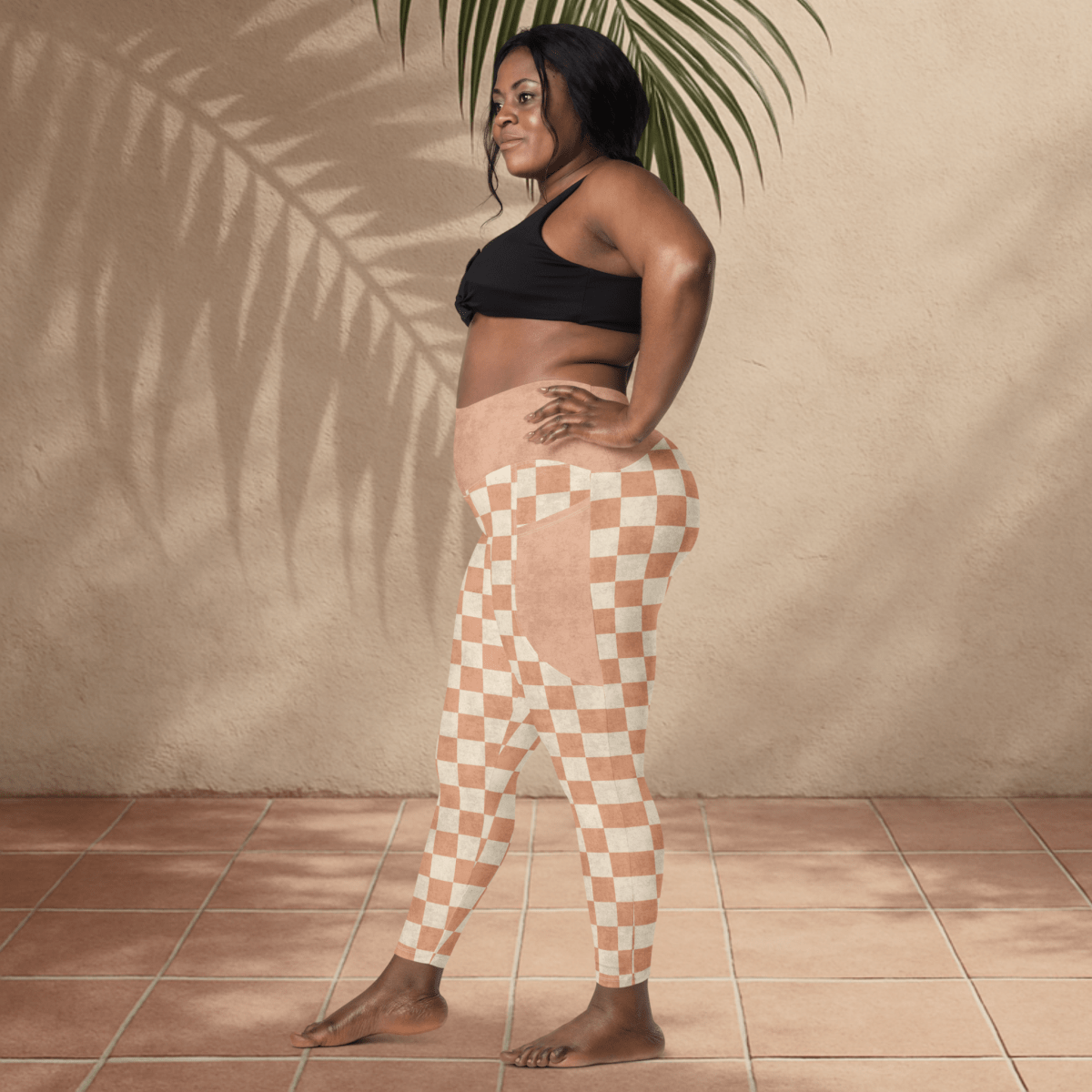 Woman wearing a black sports bra and checkered leggings standing against a beige wall with a plant.