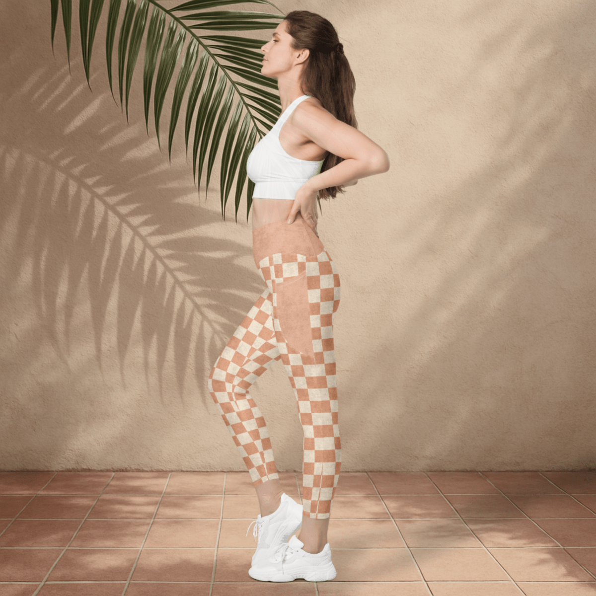 Woman wearing checkered leggings and a white sports bra against a beige wall with a palm leaf.