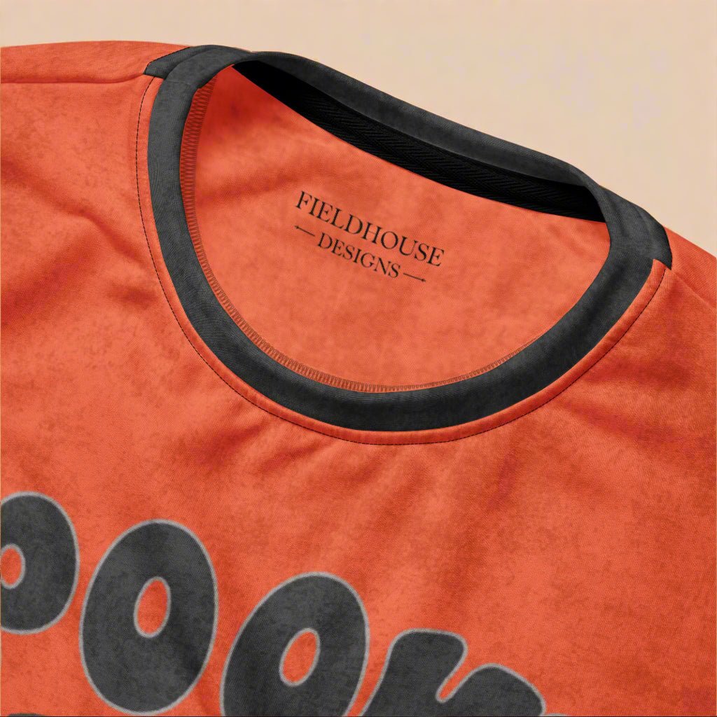 Orange shirt with black collar and 'Fieldhouse Designs' branding on a beige background