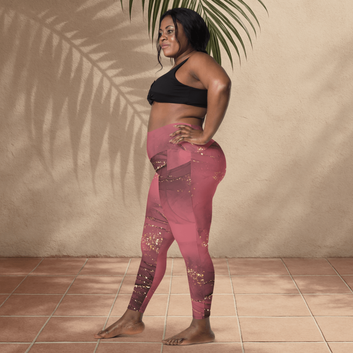 Woman wearing pink leggings and black sports bra standing against a beige wall with a plant.