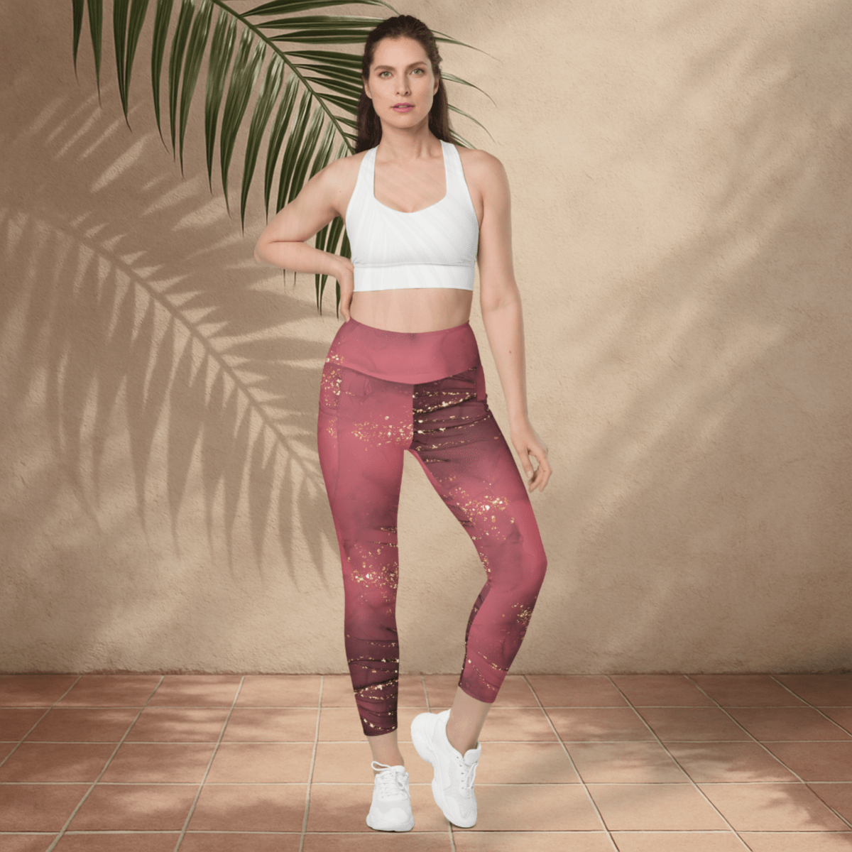 Woman wearing a white sports bra and dusty rose marble patterned leggings standing against a beige wall with a plant.